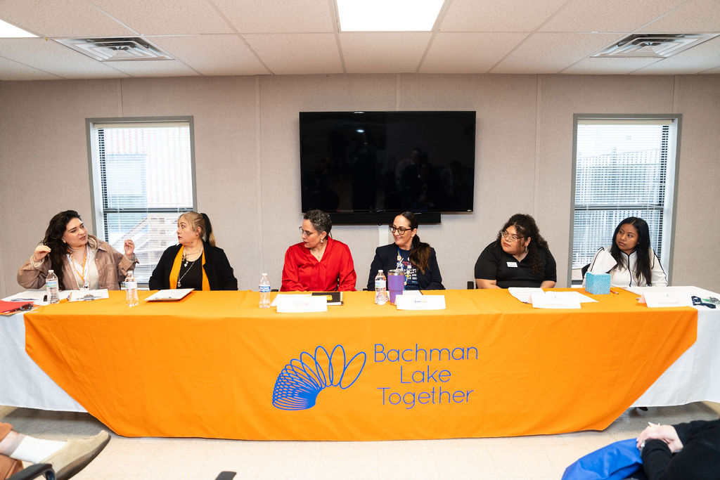 A group of six women sit together on a panel behind a table that's covered in a branded tablecloth for the nonprofit Bachman Lake Together.