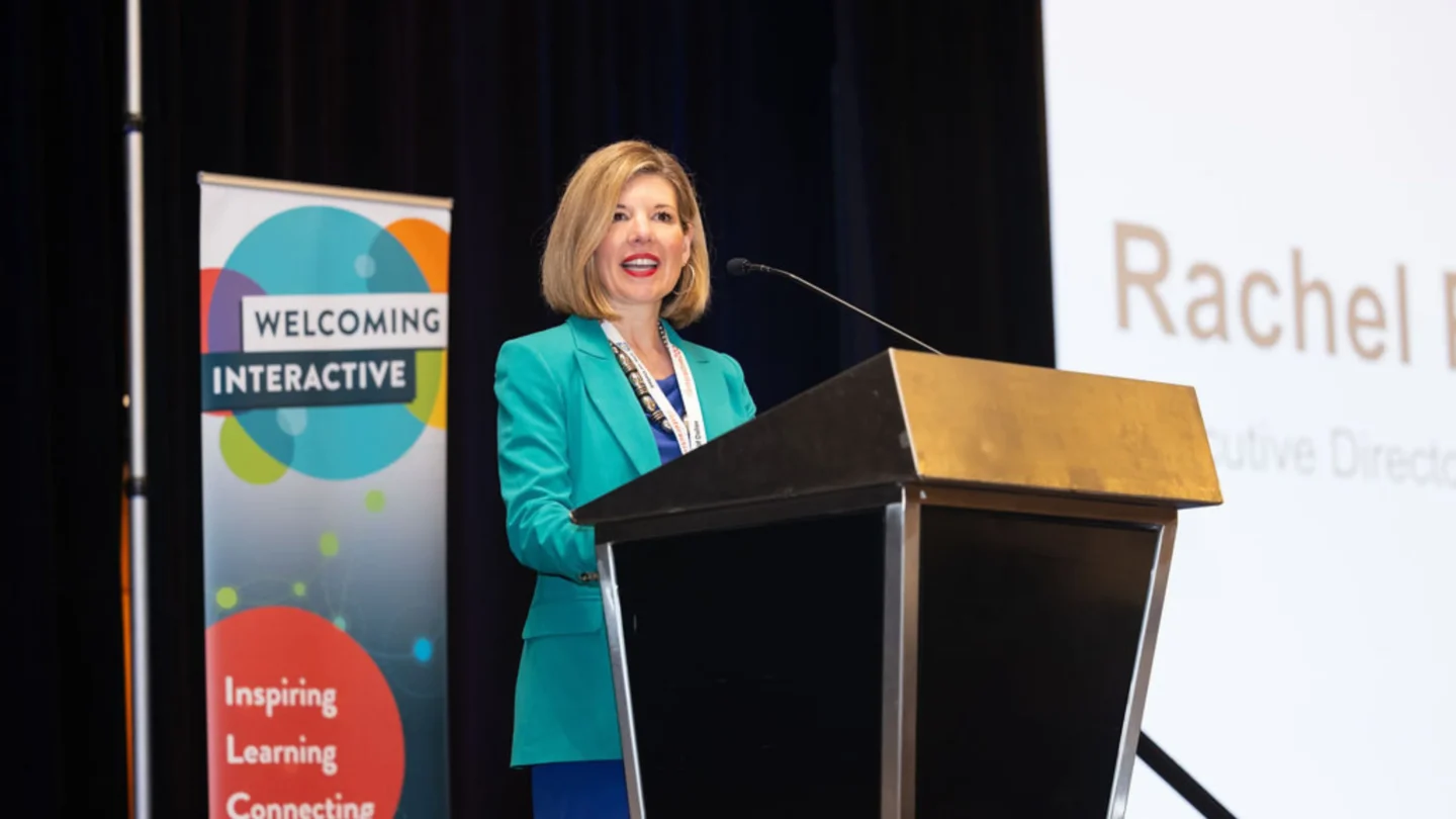 Rachel Perić stands on stage behind a lectern, presenting remarks at the Welcoming Interactive 2024.