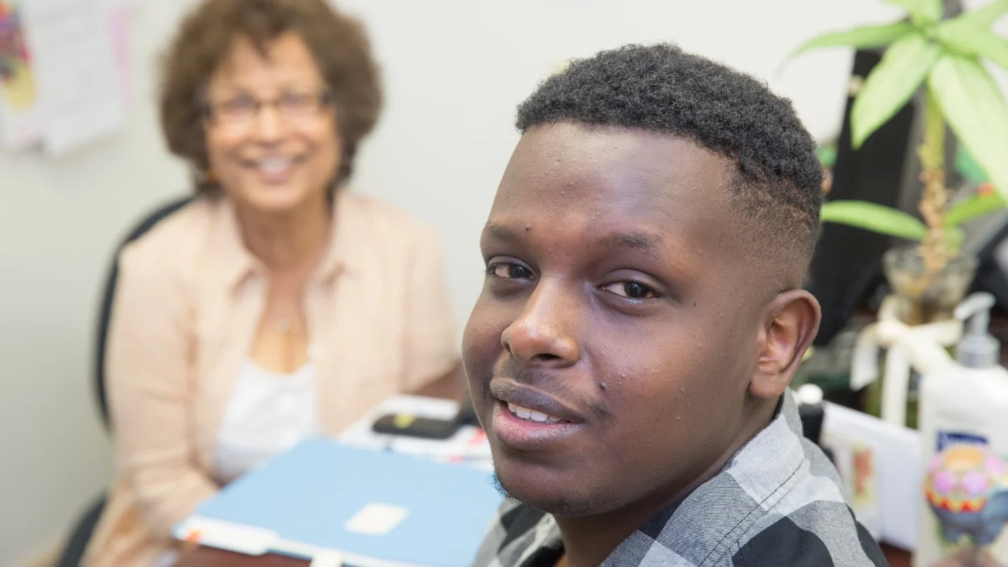 Young man smiles at the camera as a woman smiles out of focus in the background